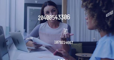 Two businesswomen discussing documents at modern office desk with data overlay