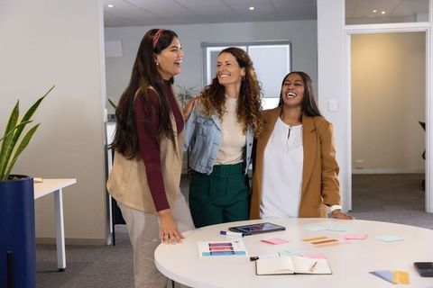 Diverse female coworkers laughing and collaborating at meeting table in modern office