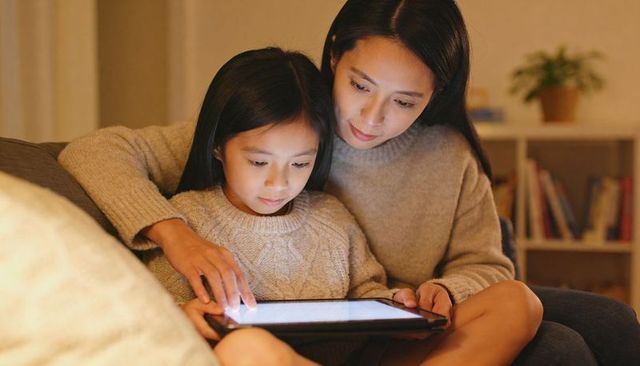 Asian mother guiding daughter on sofa with tablet glow cozy evening learning moment