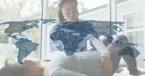 Kneeling therapist guiding patient on mat in home clinic with global health data overlay