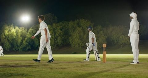 Night cricket match under floodlights with bowler, helmeted batter and umpire