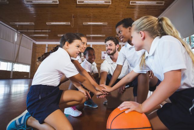 Group of school children and their sports teacher forming a hand stack in a gym. The children are wearing sports uniforms and are gathered around a basketball. This image is ideal for illustrating teamwork, sportsmanship, and physical education in schools. It can be used for educational materials, sports programs, and promotional content for school activities.