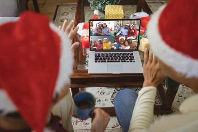 Diverse couple, seated with Santa hats, video calling a group of happy friends on a laptop during a Christmas celebration. Surrounding the laptop are festive elements like red candles and Christmas decorations. Ideal for usage in marketing materials about digital communication, holiday greetings, family gatherings, and festive celebrations.