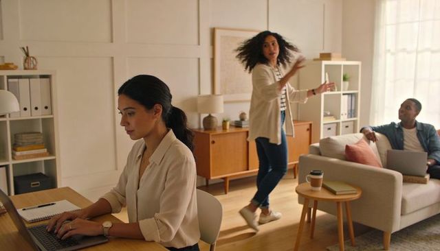 Hispanic woman typing at home desk while colleague gesturing in modern living room workspace