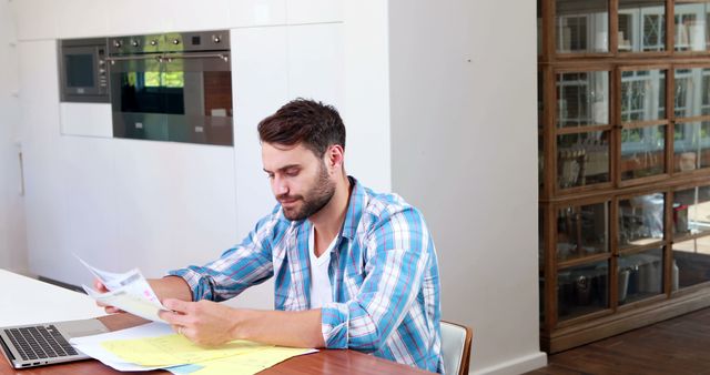 Young man reviewing paperwork while sitting at a table in a modern home office. Surroundings include sleek, minimalistic design elements and a laptop, highlighting a work-from-home setting. Ideal for themes related to remote work, home office setup, modern interiors, professional lifestyle, or productivity.