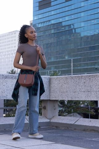 Standing on urban rooftop wearing jeans flannel tied at waist carrying brown crossbody bag