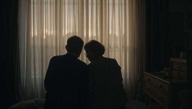 Senior couple sitting on bed leaning heads together backlit by sheer curtains morning light