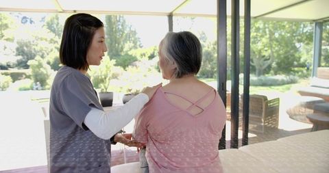 Caregiver comforting senior woman by sunlit glass doors, home care nurse aide support