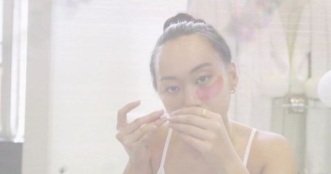 Midlife woman peeling and applying pink hydrogel eye patches at vanity during skincare routine