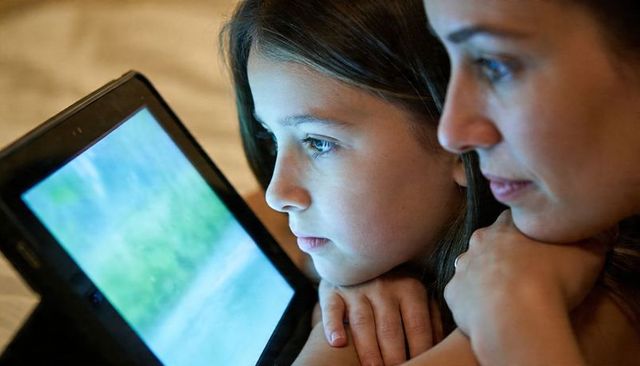 Mother and daughter cuddling on bed watching tablet glow while resting chins on hands