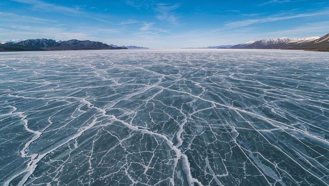 Aerial frozen lake with spiderweb ice cracks stretching to snowy mountain horizon