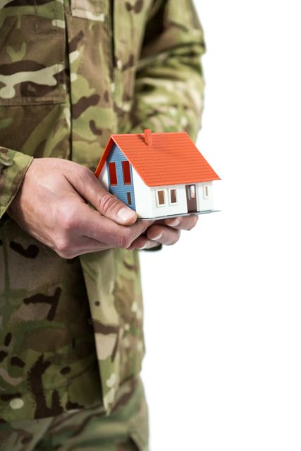 Soldier in camouflage uniform holding a small model house. Useful for themes related to military housing, homeownership, real estate for veterans, family support, and relocation assistance for military personnel.