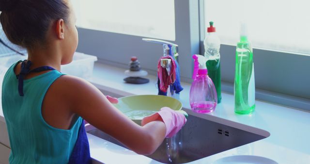 Young girl washing dishes by kitchen sink. She wears pink gloves and apron while cleaning plates. This image can be used for parenting blogs, articles on household chores, education materials on responsibility, or ads related to home cleaning products.