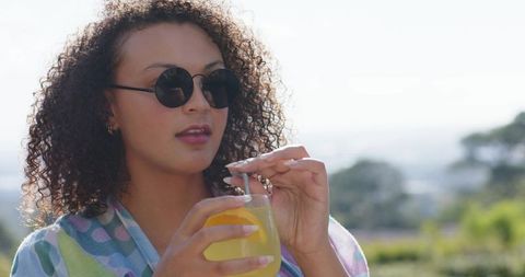Curly african american woman sipping citrus drink on sunny terrace in sunglasses and pastel shawl