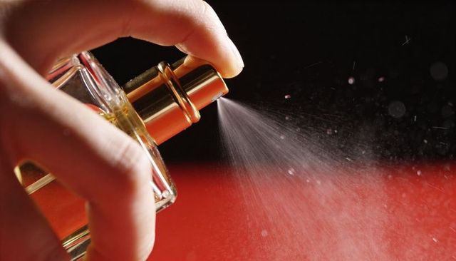 Hand pressing gold atomizer spraying luxury perfume bottle with fine mist on red backdrop