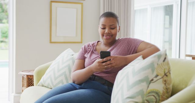 Young woman is sitting on a cozy sofa in a modern living room, smiling while using a smartphone. Ideal for themes about modern home life, casual relaxation, connectivity, and everyday technology use. Perfect for articles, blogs, and advertisements focusing on home living, comfort, and digital lifestyles.