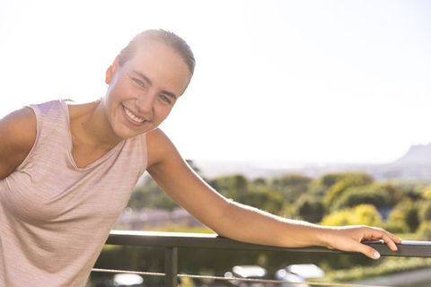 Sunlit young woman leaning on balcony railing smiling in sleeveless pink top backlit terrace