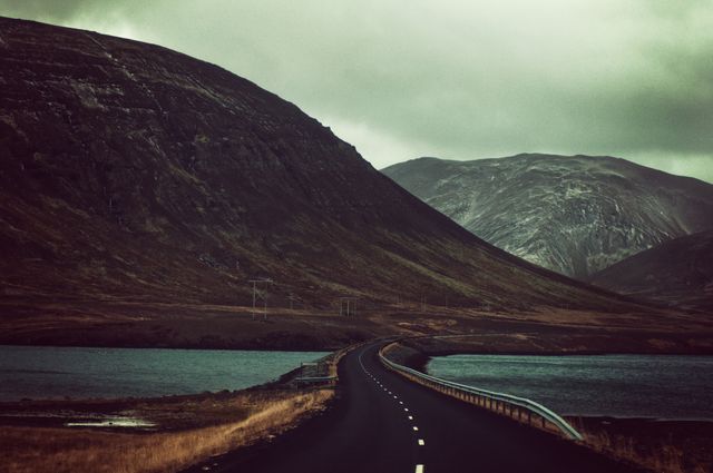 This image captures a solitary road winding between mountains and lakes during dusk, creating a dramatic and tranquil scene. Ideal for travel blogs, adventure websites, and print media promoting road trips or scenic landscapes.