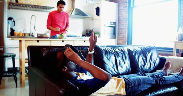 Two friends enjoying leisure time in modern home. One friend cooking in kitchen while other lying on couch using VR headset. Ideal for promoting technology in home settings, lifestyle blogs, and advertisements for home appliances or virtual reality devices.