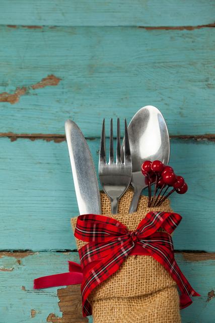 This image features a rustic table setting with cutlery wrapped in burlap and tied with a red ribbon, placed on a weathered wooden table. Ideal for use in holiday-themed content, dining and table setting articles, or rustic and vintage decor promotions.