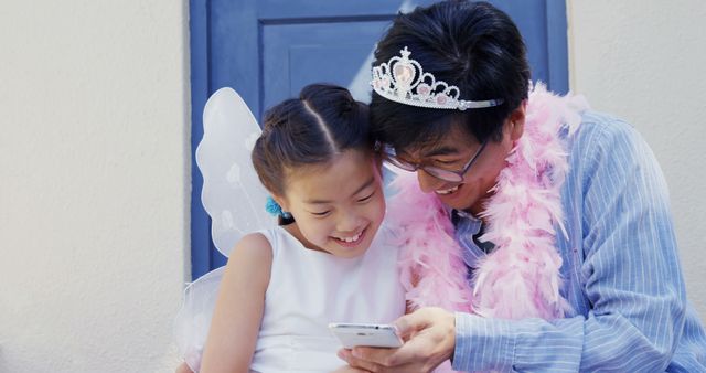 An Asian man and a young girl dressed in a fairy costume share a joyful moment looking at a smartphone, with copy space. Their laughter and costumes suggest a playful and happy family interaction.