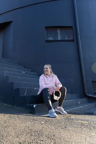Smiling woman sitting on urban steps in pink activewear holding headphones on sunlit street