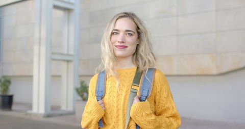 Smiling female student wearing yellow sweater holding backpack on campus walkway