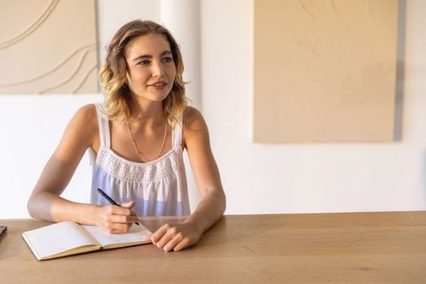Young woman journaling at sunlit wooden table in minimalist studio, lifestyle portrait