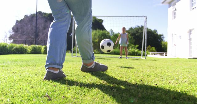 A person is kicking a soccer ball towards a goal where a child stands as a goalkeeper, with copy space. Capturing a leisurely moment, the image evokes a sense of playfulness and family bonding in an outdoor setting.