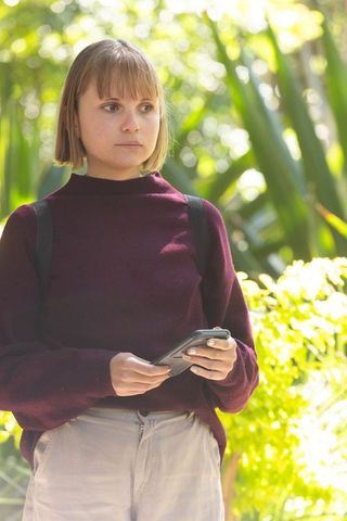 Woman standing in sunlit garden holding smartphone wearing maroon sweater and backpack