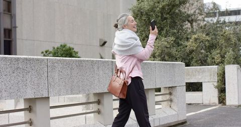 Mature woman walking urban walkway holding smartphone smiling during video call wearing scarf