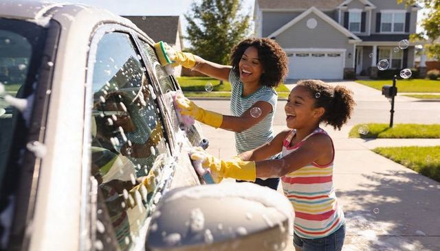 Mom and daughter washing car together on sunny suburban driveway, laughing and soapy