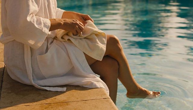 Sitting woman in linen robe dipping foot into pool water on sunlit stone deck, relaxing