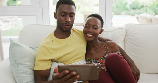 Happy african american couple sitting on sofa embracing and talking looking at tablet. staying at home in isolation during quarantine lockdown.