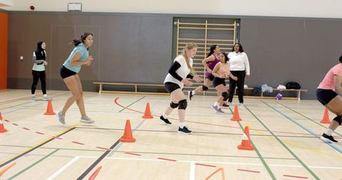 Diverse female volleyball players practicing cone agility drill in indoor gym with coach