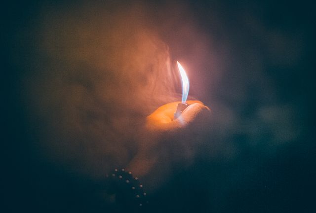 Hand holding up a lighter creates a small flame in dark, smoky surroundings. Ideal for use in projects involving themes of mystery, anxiety, dreams, spirituality, or illuminating hidden aspects.