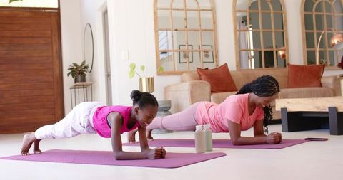 African mother and teen daughter doing forearm plank workout in sunlit living room