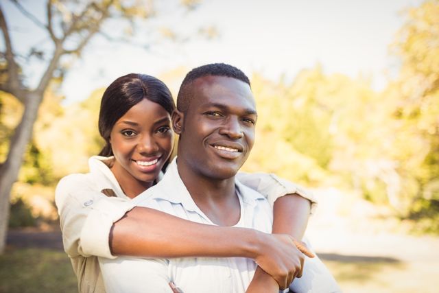 This image shows a happy African American couple embracing in a park. They are smiling and enjoying a sunny day outdoors. This image can be used for promoting relationships, love, outdoor activities, and lifestyle content.
