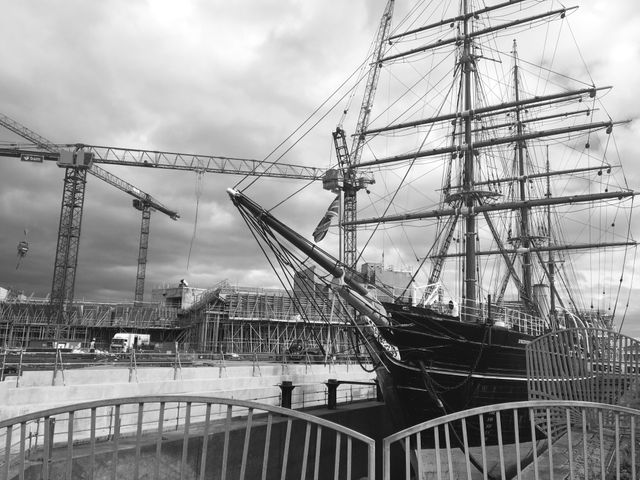 Tall ship in dock near construction site, cranes and scaffolding around, overcast sky. Useful for themes of maritime history, industrial settings, construction projects, and nautical heritage. Ideal for educational materials, documentaries, historical presentations, and industrial context designs.
