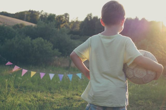 Young boy holding a soccer ball standing on a grassy field during sunset. Ideal for use in promoting outdoor activities, children's sports gear, summer camp advertisements, or childhood development programs. The serene, natural backdrop evokes feelings of freedom and joyful play.