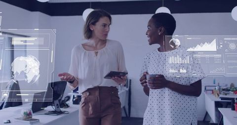 Two professional women walking through modern office discussing hud analytics and charts