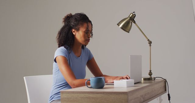 An African American woman is typing on a laptop while sitting at a modern wooden desk. The desk is minimalistic, with a straightforward design that includes a blue mug, an envelope, and a single desk lamp. She is working in an indoor setting, wearing a casual short-sleeve light blue shirt. This can be used in websites or advertisements focusing on remote work, freelance, modern office, productivity, and technology use.