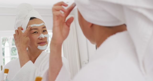 Woman standing in bathroom with white bathrobe and towel wrapped around head, applying facial mask while looking into mirror. This image can be used in articles or advertisements promoting skincare routines, beauty products, and self-care practices.