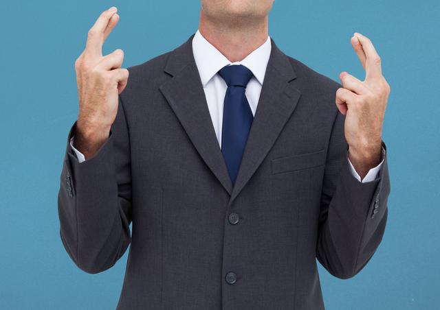 Businessman wearing suit and tie, crossing both fingers against a blue background. Ideal for themes like anticipation, good luck, corporate culture, or hopefulness in business contexts.