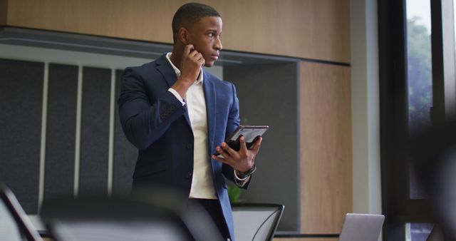 Businessman in a stylish blue suit is using a tablet device while standing in a modern office environment. Appears deep in thought. Ideal for corporate, business consultancy, technology, and professional services related content and websites.