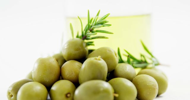 This image shows a heap of green olives with a sprig of rosemary and a glass of olive oil in the background, all on a white surface. Ideal for use in health and food blogs, Mediterranean cuisine recipes, cooking websites, and promotional materials for organic food products.