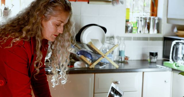 Middle-aged woman using laptop in kitchen with curly hair and casual attire - Download Free Stock Images Pikwizard.com