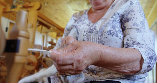 Senior Caucasian woman spins yarn on an old-fashioned spinning wheel at home. Her skilled hands craft homemade textiles, showcasing a traditional hobby.