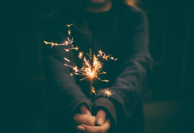 Person holding sparkler in outstretched hands, creating small fireworks effect in dark environment. Can be used for festive occasions, celebratory posts, or New Year celebrations.