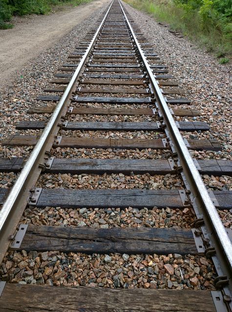 View of railroad tracks stretching into distance with green vegetation on both sides. Useful for topics on transport, travel, journeys, and infrastructure.